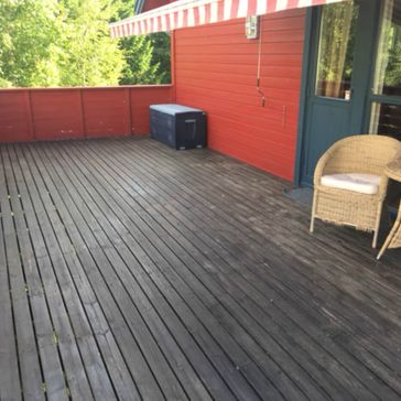 Gammel terrasse med svartmalt gulv og rødt gelender med persienner på vegg og utemøblemang 